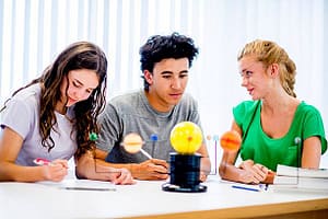 A multi-ethnic group of college age students are sitting together in astronomy class studying a model of our solar system, while working on a homework assignment.